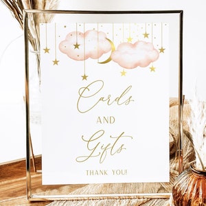 Puede incluir: Un cartel blanco con letras doradas en un marco dorado. El cartel dice "Tarjetas y regalos ¡Gracias!" y tiene un diseño de nubes y estrellas rosas y doradas.