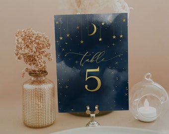 May include: A navy blue table number card with gold stars and a crescent moon. The card says "table 5" in gold lettering. The card is on a gold metal stand.