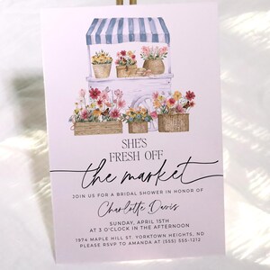 Op de afbeelding: Een roze bruidsdouche-uitnodiging met een aquarel-illustratie van een bloemenkraam. De tekst luidt "She's Fresh Off the Market" en bevat de naam van de bruid, datum, tijd en locatie.