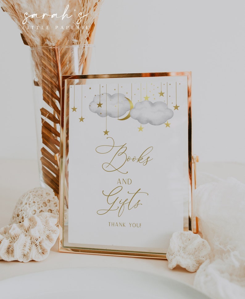 May include: A gold framed sign with a white background and gold lettering. The sign says "Books and Gifts" with a crescent moon and stars graphic above the text. The sign is sitting on a table with a white surface.