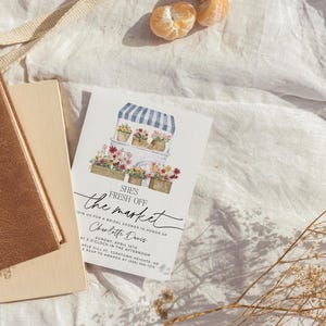 Op de afbeelding: Een aquarel-illustratie van een bloemenkar met een blauw-wit gestreept luifel. De tekst "SHE'S FRESH OFF THE MARKET" is geschreven in zwarte cursieve letters. De uitnodiging is voor een bruidsdouche ter ere van Charlotte Davis op zondag 15 april om 15:00 uur. Het adres is Aple Hill St. Yorktown Heights, NY. RSVP aan Amanda op (555) 555-1212.