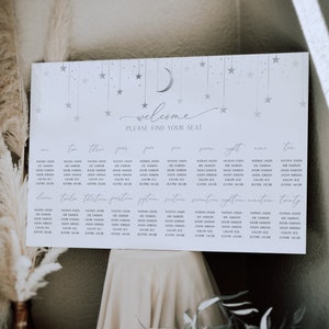 May include: A white seating chart with a silver crescent moon and stars. The chart is titled "Welcome, Please Find Your Seat." The chart lists names for each table number, from one to twenty.