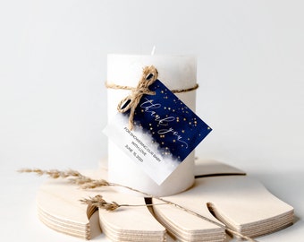 Può includere: Una candela a colonna bianca con un fiocco di spago e un'etichetta blu navy. L'etichetta ha stelle dorate e le parole "grazie" e "Per aver festeggiato il nostro bambino con amore 15 giugno 2020."