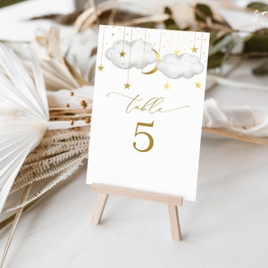 May include: White table number card with gold lettering that says "table 5" and a gold crescent moon and clouds with gold stars hanging from them.