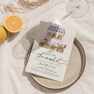 Op de afbeelding: Een witte kaart met een bloemillustratie van een bloemenkraam met een blauw-wit gestreept luifel. De tekst "SHE'S FRESH OFF the market" is geschreven in zwarte cursieve letters. De tekst "JOIN US FOR A BRIDAL SHOWER IN HONOR OF Charlotte Davis" is geschreven in zwarte tekst. De tekst "SUNDAY, APRIL 15TH AT 3 O'CLOCK IN THE AFTERNOON 1974 MAPLE MILL ST. YORKTOWN HEIGHTS, NO PLEASE RSVP TO AMANDA AT (555) 555-1212" is geschreven in zwarte tekst.