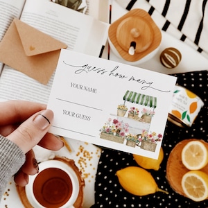May include: A white card with the text "guess how many" in black cursive writing. There are two blank lines for "your name" and "your guess". The card features a watercolor illustration of a white flower cart with green awning and flowers in pots.