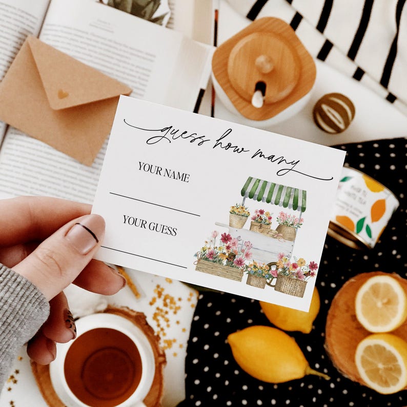 May include: A white card with the text "guess how many" in black cursive writing. There are two blank lines for "your name" and "your guess". The card features a watercolor illustration of a white flower cart with green awning and flowers in pots.