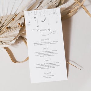 May include: A white menu card with a silver crescent moon and stars design. The menu features appetizers, salad, entree, and dessert options. The text reads: "meny", "Appetizer", "Sun-dried Tomato Risotto Cakes with Basil Aioli", "Crab Bisque with Aioli and Garlic Croutons", "Salad", "Mesclun Greens with Pears and Spiced Walnuts in Sherry Vinaigrette", "Mixed Greens with Crispy Duck in Elder Orange Vinaigrette", "Entree", "Sesame Seed Crusted Seared Tuna Steak with Five Spice Braised Chard", "Wild Mushroom Strudel with Roasted Red Peppers and Goat Cheese", "Dessert", "Coconut Layer Cake", "Lime Gelato"