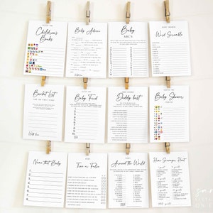 Puede incluir: Un conjunto de tarjetas de juegos para baby shower, con t&iacute;tulos como "Adivina los libros infantiles" y "Consejos para el beb&eacute;". Las tarjetas est&aacute;n impresas en tinta negra sobre papel blanco y sujetas con pinzas de madera.