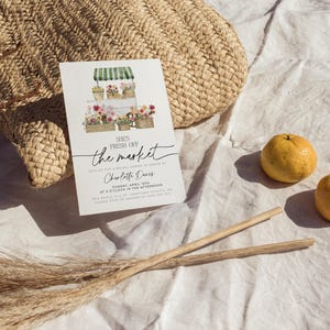 Pu&ograve; includere: Un biglietto d'invito bianco con il testo "She's Fresh Off the Market" e illustrazioni floreali. Il biglietto &egrave; appoggiato su una borsa di paglia intrecciata e tessuto bianco, con arance e bastoncini di legno nelle vicinanze. L'invito &egrave; per un addio al nubilato.
