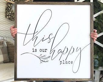 May include: A white wooden sign with black calligraphy that reads "This is our happy place" with a small floral design. The sign is being held by a person.