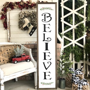 May include: A white wooden sign with a black painted word "BELIEVE" in a decorative font. The sign is framed with a brown wood frame and has a green sprig with red berries at the top and bottom.