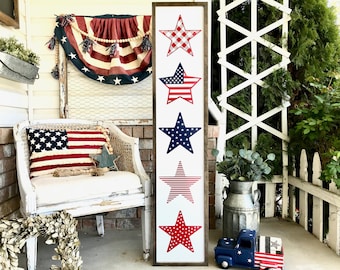 May include: A wooden sign with five red, white, and blue stars. The stars are in a vertical row and are decorated with different patterns. The sign is hanging on a white porch.