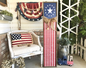 May include: A wooden sign featuring a red, white and blue American flag design. The sign is decorated with a hessian bow and is leaning against a white chair on a porch.