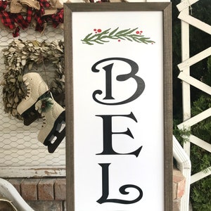May include: A white wooden sign with a brown frame, featuring the word "BEL" in black stylized lettering. A green sprig with red berries is at the top of the sign.