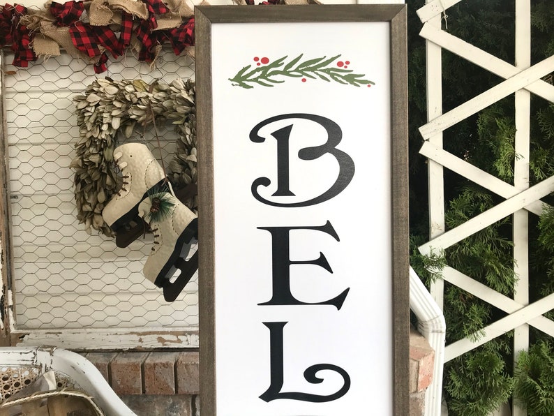 May include: A white wooden sign with a brown frame, featuring the word "BEL" in black stylized lettering. A green sprig with red berries is at the top of the sign.