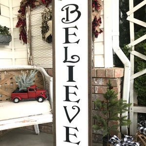 May include: A white wooden sign with the word "BELIEVE" written in black cursive letters. The sign is decorated with green pine branches and red berries. The sign is leaning against a white brick wall.