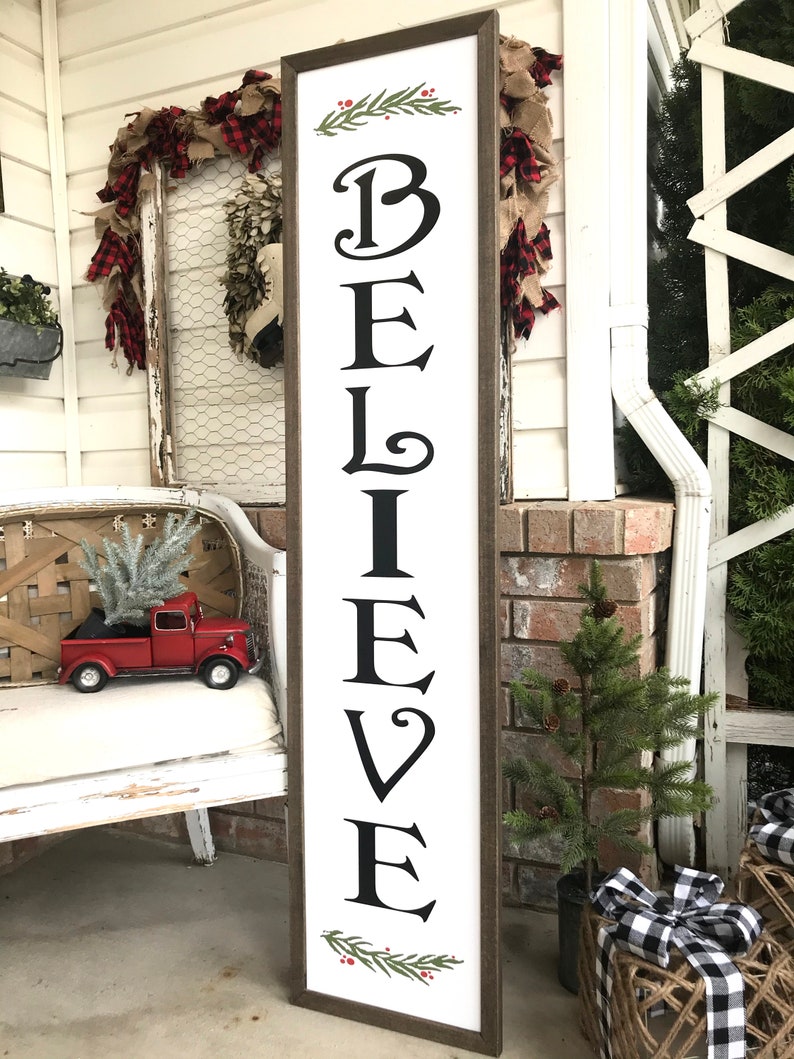May include: A white wooden sign with the word "BELIEVE" written in black cursive letters. The sign is decorated with green pine branches and red berries. The sign is leaning against a white brick wall.