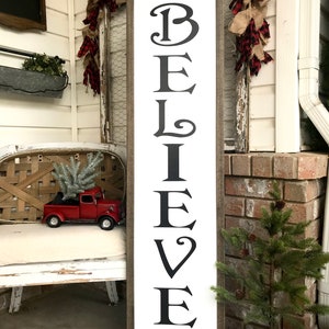 May include: A white wooden sign with the word "BELIEVE" in black lettering. The sign is framed in brown wood and has a green garland with red berries at the top and bottom.