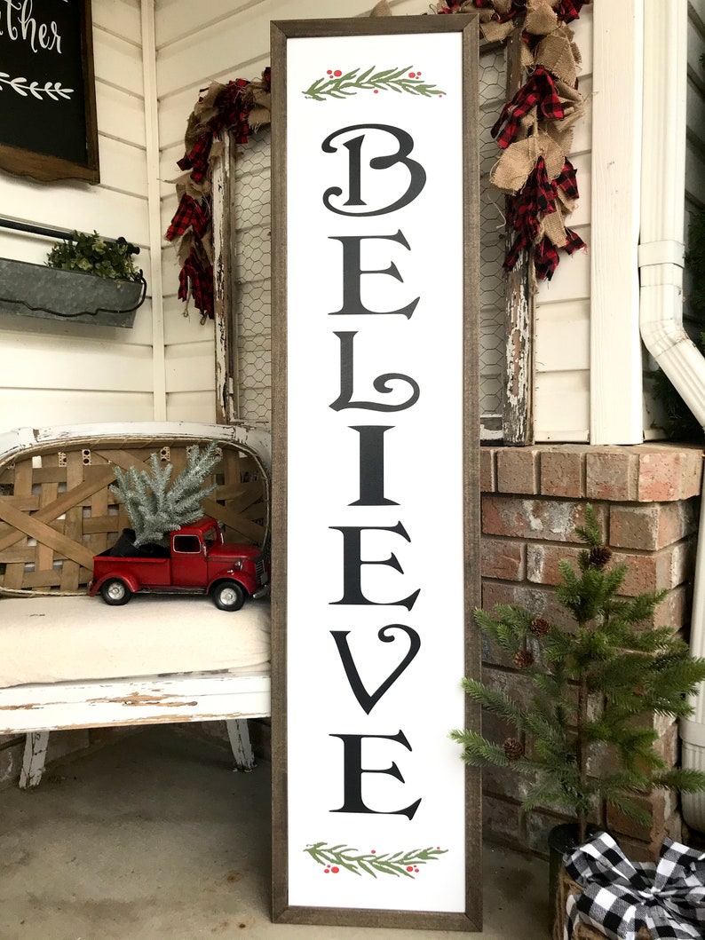 May include: A white wooden sign with the word "BELIEVE" in black lettering. The sign is framed in brown wood and has a green garland with red berries at the top and bottom.