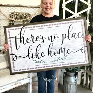 May include: A wooden sign with a white background and a brown frame. The sign has a white and brown woodgrain pattern. The sign says "there's no place like home" in black cursive lettering with a green floral design underneath the text. The sign is surrounded by a brown border with a row of small purple dots.