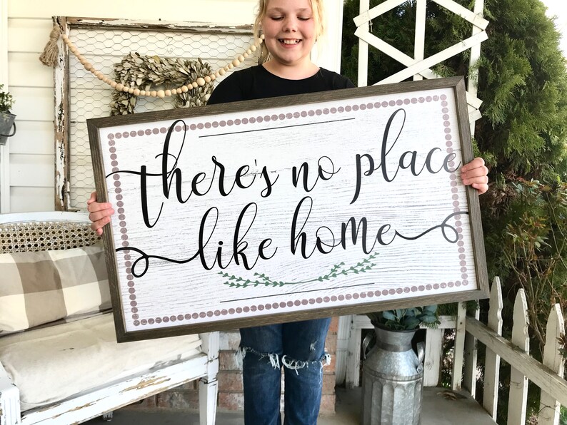 May include: A wooden sign with a white background and a brown frame. The sign has a white and brown woodgrain pattern. The sign says "there's no place like home" in black cursive lettering with a green floral design underneath the text. The sign is surrounded by a brown border with a row of small purple dots.