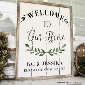 K&ouml;nnte beinhalten: Ein wei&szlig;es Holzschild mit schwarzem Text, der "Willkommen in unserem Zuhause" lautet, mit einem gr&uuml;nen Blattkr&auml;nzchen-Design. Das Schild enth&auml;lt auch die Namen "KC & Jessika" und "Elsa Leith Noah Carter". Das Schild ist in einem hellbraunen Holzrahmen gerahmt.