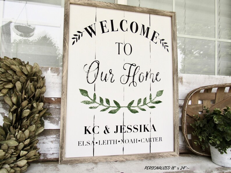 K&ouml;nnte beinhalten: Ein wei&szlig;es Holzschild mit schwarzem Text, der "Willkommen in unserem Zuhause" lautet, mit einem gr&uuml;nen Blattkr&auml;nzchen-Design. Das Schild enth&auml;lt auch die Namen "KC & Jessika" und "Elsa Leith Noah Carter". Das Schild ist in einem hellbraunen Holzrahmen gerahmt.