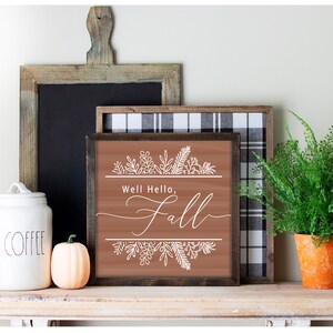 May include: A brown wooden sign with the text "Well Hello, Fall" in white script. The sign is decorated with white floral designs and has a rustic wood frame.