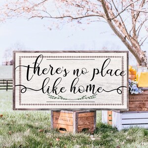 May include: A wooden sign with a white background and brown border. The sign says "There's no place like home" in black cursive lettering with green leaves on either side of the word "home".