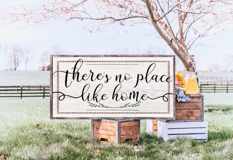 May include: A wooden sign with a white background and brown border. The sign says "There's no place like home" in black cursive lettering with green leaves on either side of the word "home".