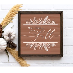 May include: A brown wooden framed sign with the text "Well Hello, Fall" in white cursive font. The sign is decorated with white floral designs at the top and bottom.