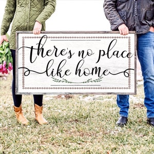 May include: A couple hold a wooden sign with the text "There's no place like home" in black cursive lettering. The sign has a white background with a brown border and a decorative pattern of small circles. The couple are standing in a field with green grass and some snow on the ground.