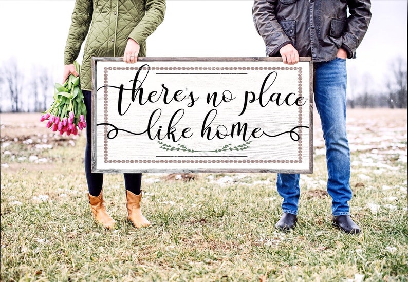 May include: A couple hold a wooden sign with the text "There's no place like home" in black cursive lettering. The sign has a white background with a brown border and a decorative pattern of small circles. The couple are standing in a field with green grass and some snow on the ground.