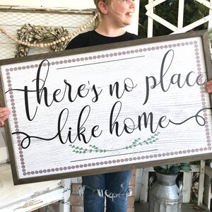May include: A wooden sign with a white background and a brown frame. The sign has a pink and white polka dot border and says "there's no place like home" in black cursive lettering with green leaves underneath the text.