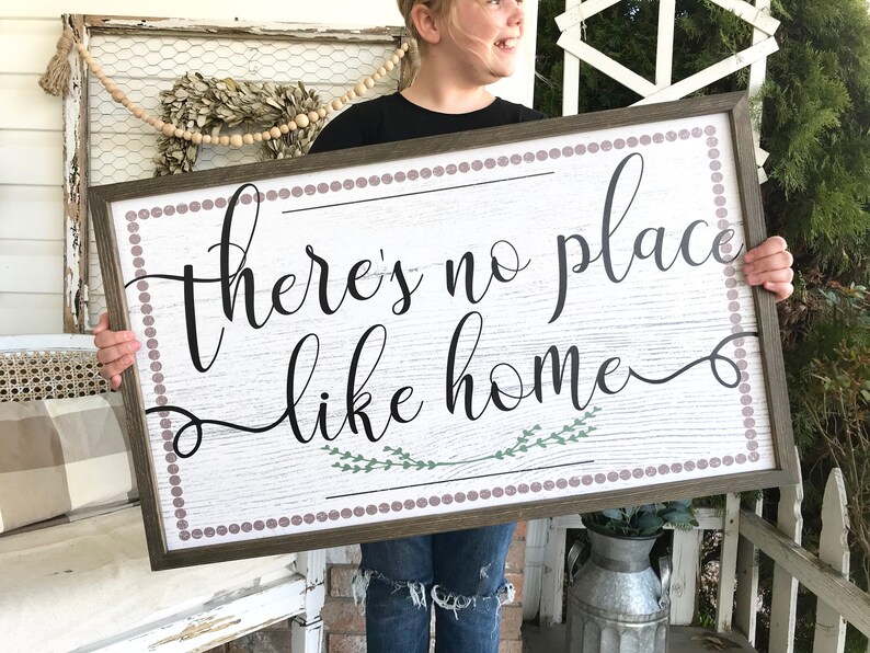 May include: A wooden sign with a white background and a brown frame. The sign has a pink and white polka dot border and says "there's no place like home" in black cursive lettering with green leaves underneath the text.