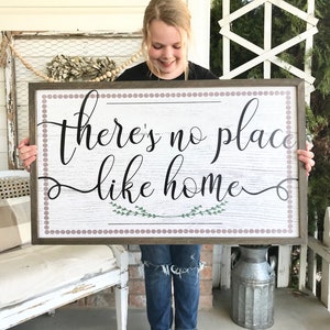 May include: A wooden sign with a whitewashed background and a brown frame. The sign has a pink and white polka dot border and says "there's no place like home" in black cursive lettering. There are two small green sprigs of leaves on either side of the word "home".