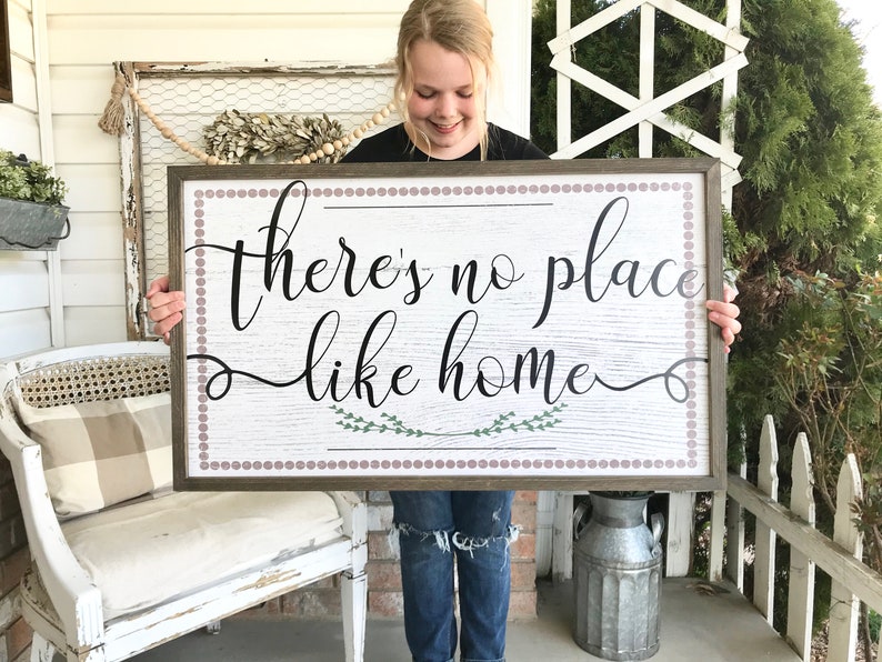 May include: A wooden sign with a whitewashed background and a brown frame. The sign has a pink and white polka dot border and says "there's no place like home" in black cursive lettering. There are two small green sprigs of leaves on either side of the word "home".