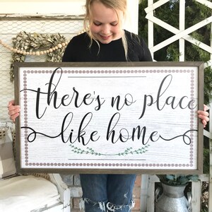 May include: A wooden sign with a white background and brown frame. The sign has a brown and white polka dot border and says "there's no place like home" in black cursive lettering with green leaves underneath the text.