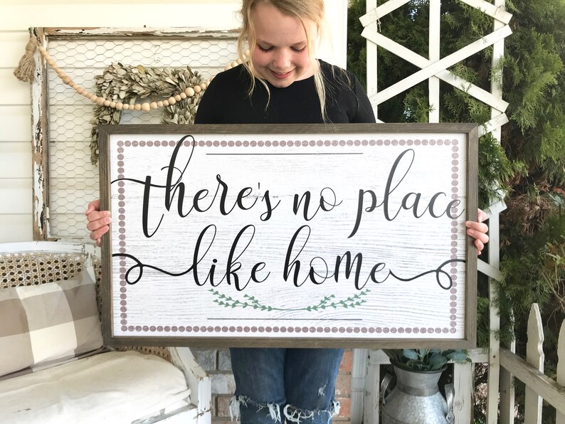 May include: A wooden sign with a white background and brown frame. The sign has a brown and white polka dot border and says "there's no place like home" in black cursive lettering with green leaves underneath the text.