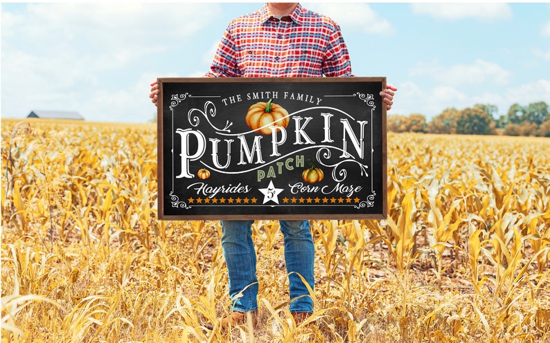 May include: A black chalkboard sign with white text that reads "The Smith Family Pumpkin Patch Hayrides Corn Maze". The sign is decorated with a pumpkin and stars. A person is holding the sign in front of a field of corn.