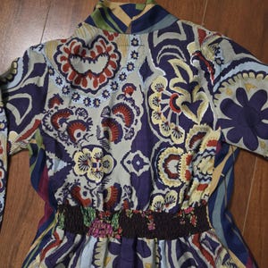 Pode incluir: Uma camisa de manga comprida com um padr&atilde;o floral e paisley multicolorido vibrante em tons de roxo profundo, azul claro, creme e vermelho ferrugem. A pe&ccedil;a inclui uma cintura el&aacute;stica franzida castanho-escura e uma gola alta.