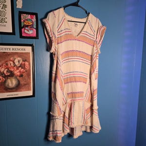May include: Cream-colored dress with horizontal stripes in pink, blue, and peach. The dress has a V-neck, short sleeves, and a ruffled hem. The fabric appears to be textured, with a frayed edge detail. The dress is hanging on a black hanger.