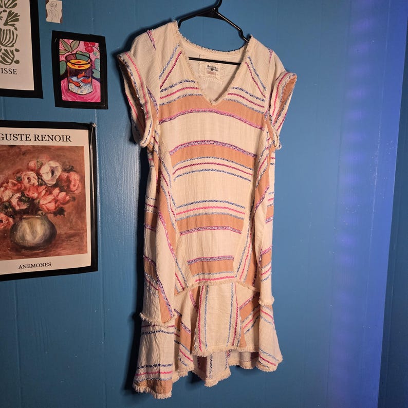 May include: Cream-colored dress with horizontal stripes in pink, blue, and peach. The dress has a V-neck, short sleeves, and a ruffled hem. The fabric appears to be textured, with a frayed edge detail. The dress is hanging on a black hanger.