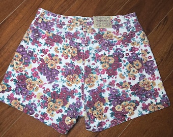 Puede incluir: Pantalones cortos vaqueros de cintura alta con un vibrante estampado floral. Los pantalones cortos presentan una base blanca cubierta con una mezcla de flores moradas, burdeos y amarillas. Los bolsillos traseros y la cintura son visibles.