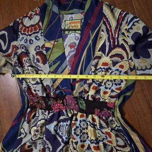 Pode incluir: Um vestido da marca Johnny Was com decote em V e cintura franzida, apresentando um padr&atilde;o floral e geom&eacute;trico multicolorido em tons de azul, vermelho e creme. Uma fita m&eacute;trica amarela sobre o peito mostra uma largura de cerca de 43 cent&iacute;metros.