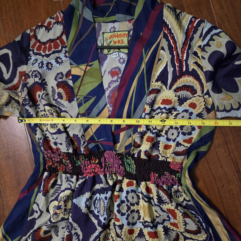 Pode incluir: Um vestido da marca Johnny Was com decote em V e cintura franzida, apresentando um padr&atilde;o floral e geom&eacute;trico multicolorido em tons de azul, vermelho e creme. Uma fita m&eacute;trica amarela sobre o peito mostra uma largura de cerca de 43 cent&iacute;metros.