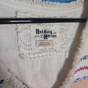 May include: Close-up of a white top with fringe detailing and a crochet neckline. The tag reads "Holding Horses" and "Made in India". The top has pink and blue embroidered stripes.