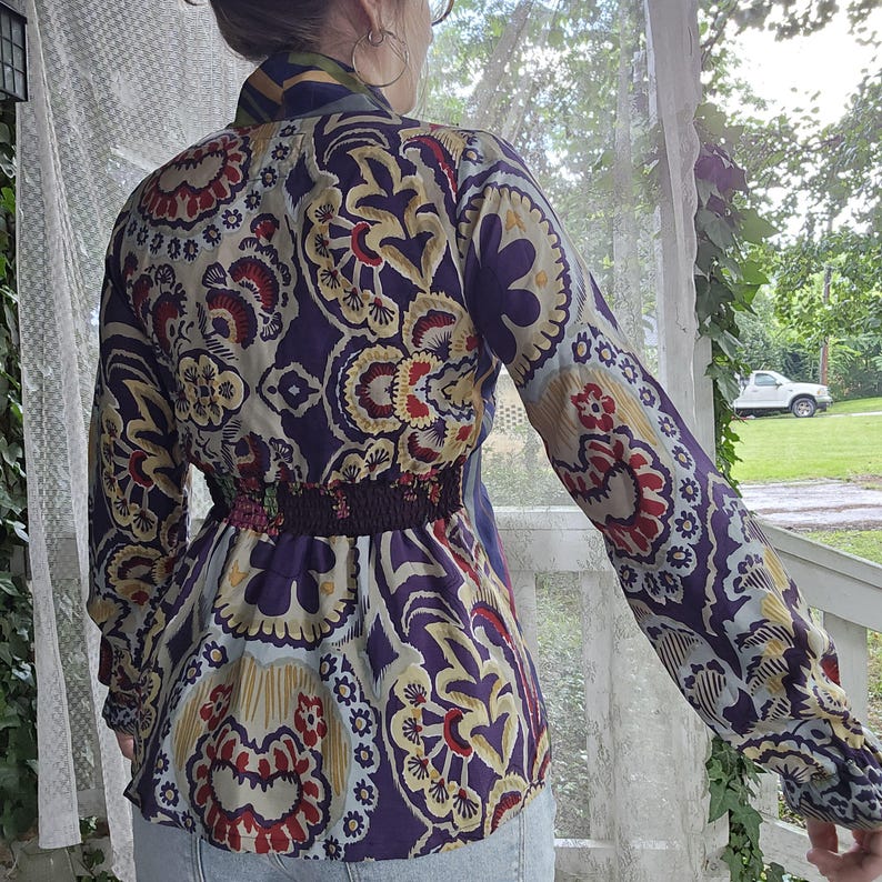 Pode incluir: Uma blusa de manga comprida com um padr&atilde;o vibrante e intrincado de paisley e flores em tons de roxo profundo, creme, vermelho e dourado. A pe&ccedil;a inclui um detalhe de cintura el&aacute;stica franzida e uma gola alta, exibindo um design ousado.