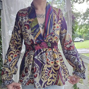 Pode incluir: Uma blusa de manga comprida com decote em V profundo e cintura el&aacute;stica franzida. A pe&ccedil;a apresenta um padr&atilde;o floral e paisley multicolorido vibrante em tons de roxo, dourado, vermelho e creme, com mangas compridas e punhos.
